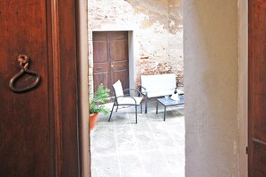 Una porta in legno aperta che dà su un cortile con un muro in mattoni, una pianta in vaso, una sedia bianca e un tavolo con un bicchiere.