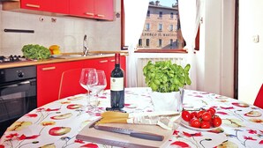 Ett kök med röda skåp, ett bord med ett blommigt bordsduk, en vinflaska, glas, en skärbräda, en kniv, en skål med tomater och en basilikaplant.
