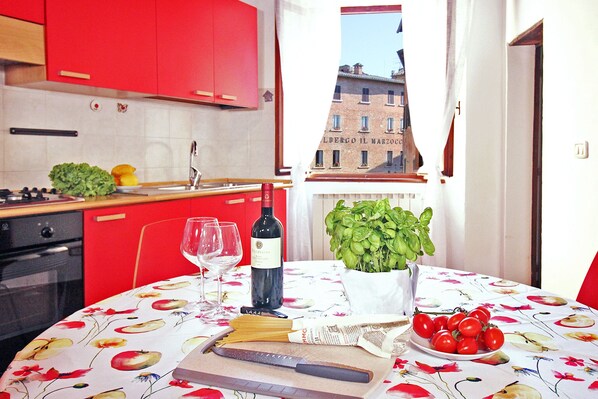 Una cucina con mobili rossi, un tavolo con tovaglia floreale, una bottiglia di vino, bicchieri, un tagliere, un coltello, una ciotola di pomodori e una pianta di basilico.