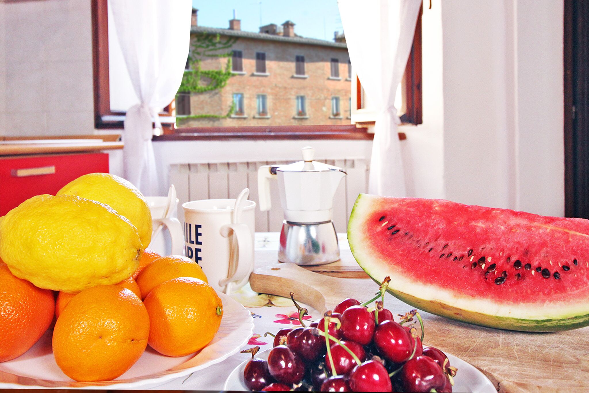 A kitchen counter with a plate of oranges, a bowl of cherries, a sliced watermelon, and a coffee maker.