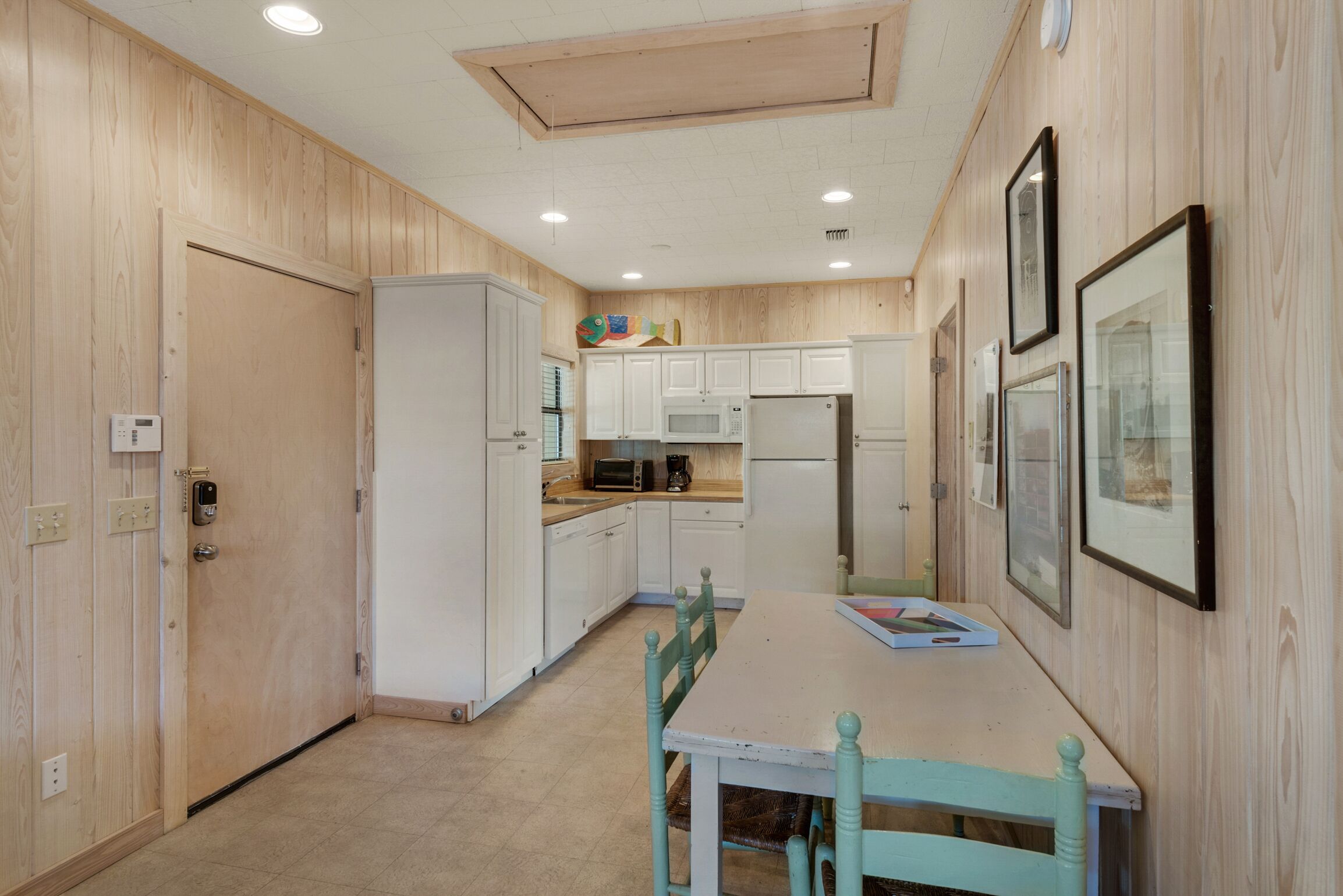 A compact kitchen with white cabinets, a microwave, and a refrigerator. There is a dining table with chairs and a few framed pictures on the wall.