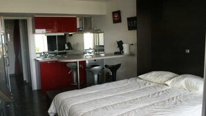 A bedroom with a large bed, a kitchenette with red cabinets, and a bathroom visible through an open door. - Air-conditioned studio with pool, parking, wifi - Cap d'Agde (Cap d'Agde naturisme)