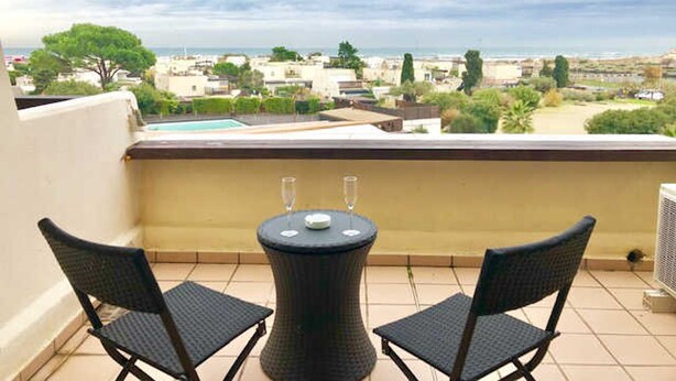 A balcony with two chairs and a table, overlooking a coastal town with buildings and a view of the sea. - Air-conditioned studio with pool, parking, wifi - Cap d'Agde (Cap d'Agde naturisme)