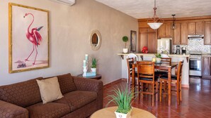 Una sala de estar con un sofá marrón, una mesa de comedor con sillas, una zona de cocina con armarios de madera y un cuadro de un flamenco en la pared.