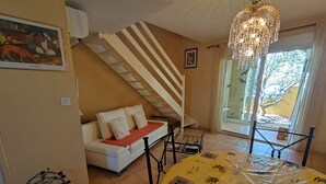 Um quarto acolhedor com um sofá branco, uma mesa coberta por um pano de mesa amarelo, uma escada e uma janela grande com vista para árvores.