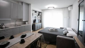 A modern hotel room with a kitchenette, a bed with a gray bedspread and striped pillows, a wooden dining table with a placemat, and a large window with sheer curtains. - Urban Zebra Modern Studio in Donizetti Residence (Bergamo)