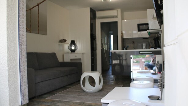 A modern living room with a grey sofa, a white coffee table, and a small white stool. There is a kitchen area with white cabinets and a microwave. - Charming Villa in Agde: 2BR, A/C, Private Terrace, WiFi, Parking (Cap d'Agde naturisme)