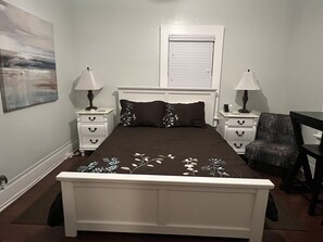 Ein Schlafzimmer mit einem weißen Bett, zwei Nachttischen mit Lampen, einem Sessel und einem Fenster mit Jalousien.