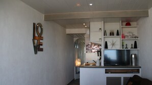 A modern living room with a sofa, a coffee table, and a TV. There is a kitchen area with a fridge, microwave, and a small island. The room has a minimalist design with white walls and a wooden ceiling. - Air-conditioned apartment for 2 in Cap d'Agde (Cap d'Agde naturisme)
