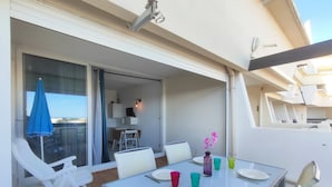 Un coin repas avec une table dressée pour deux personnes, des chaises, une petite table ornée d’un vase de fleurs et une vue sur l’intérieur à travers une porte vitrée.