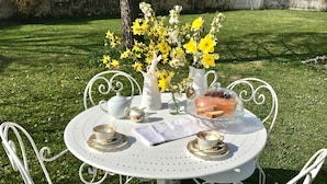 Een ronde metalen tafel met een witte tafelkleed, omringd door witte metalen stoelen. Op de tafel staat een witte karaf, een theepot, een kopje met schotel, en een vaas met gele bloemen.