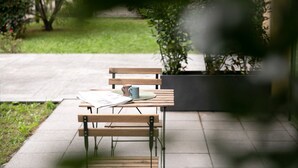 Un banc en bois avec une table, une tasse et un menu dessus, installé sur une terrasse en béton avec une zone herbeuse et un bâtiment en arrière-plan.