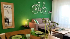 Un salon avec un mur vert, une œuvre d’art représentant un vélo, un canapé et une table à manger agrémentée de sets de table à motifs de feuilles vertes.