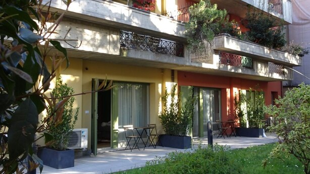 Un immeuble résidentiel à plusieurs étages, doté de balcons, d’un espace extérieur aménagé pour s’asseoir et de plantes en pot.