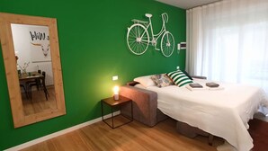 Une chambre avec un mur vert, une décoration représentant un vélo blanc, un lit recouvert d’une couverture blanche, une table de chevet avec une lampe et un miroir sur le côté gauche.