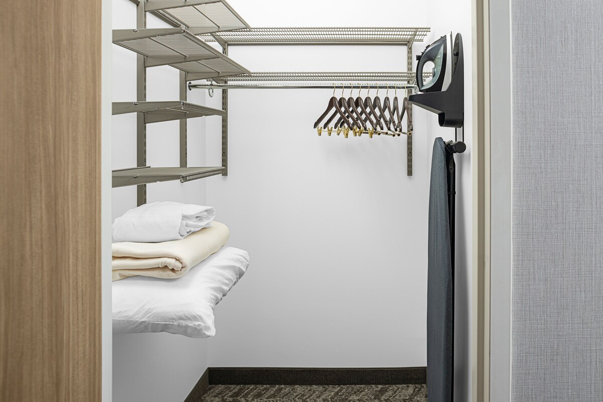 Une pièce de taille modeste, équipée d’une étagère métallique, d’un portant à vêtements et d’un lit dont le linge de lit est soigneusement plié.