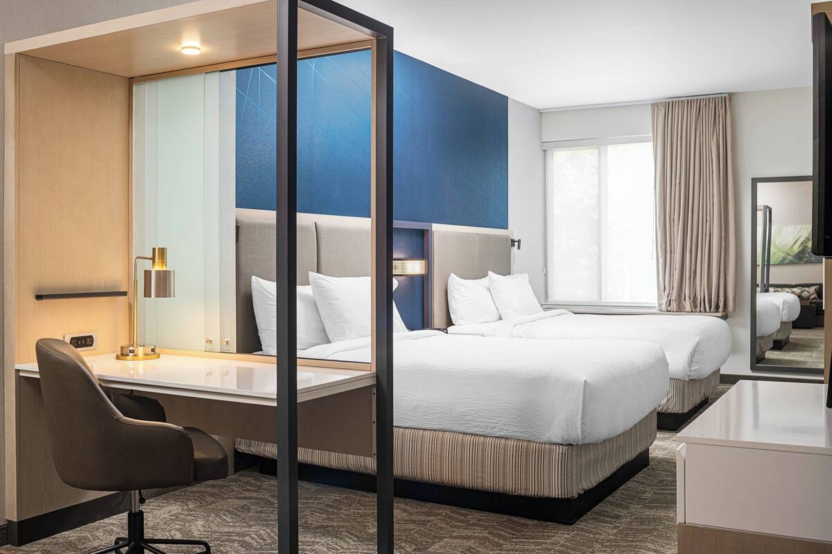 Une chambre d’hôtel équipée d’un bureau, de deux lits, d’un grand miroir et offrant une vue sur l’extérieur.