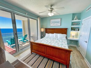 Ein Schlafzimmer mit Bett, Deckenventilator, Meerblick durch eine Schiebetür und einem kleinen Balkon mit Stühlen.