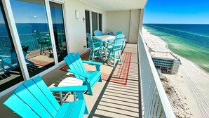 Un balcon avec des chaises turquoise et une table, donnant sur une plage aux eaux d’un bleu limpide.