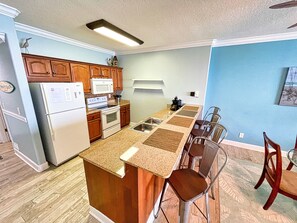 Une cuisine avec des meubles de rangement en bois, un réfrigérateur blanc, un four à micro-ondes et une cuisinière. On y trouve également un coin repas avec une table et des chaises.
