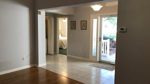 Un'ampia sala con pavimento in legno, una porta che conduce a una camera da letto e vista su un giardino attraverso una porta vetrata.