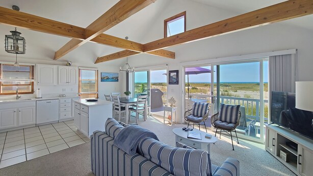 A spacious living area with a kitchen, dining table, and a balcony with a view of the beach. - Waterfront 2BD Cottage: RIGHT ON THE BEACH, Water View, Direct Beach Access, A/C & Exclusive Use Deck (North Truro)