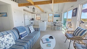 A spacious living area with a sofa, a coffee table, and a dining table. There is a kitchen area with white cabinets and a small island. The room has large windows and a balcony with a view of the beach. - Waterfront 2BD Cottage: RIGHT ON THE BEACH, Water View, Direct Beach Access, A/C & Exclusive Use Deck (North Truro)