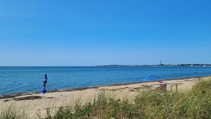 A beach with clear blue water, a sandy shore, and a distant shoreline with buildings and a lighthouse. - Waterfront 2BD Cottage: RIGHT ON THE BEACH, Water View, Direct Beach Access, A/C & Exclusive Use Deck (North Truro)