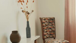 Ein Holzstuhl mit einem geometrisch gemusterten Kissen, eine Vase mit Trockenpflanzen und ein weißer Tisch mit einer braunen Vase.