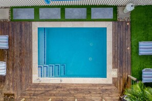 Una piscina rettangolare con una terrazza in legno tutt'intorno, una piccola area relax con una panca, e una zona sopra la piscina rivestita con erba sintetica verde.