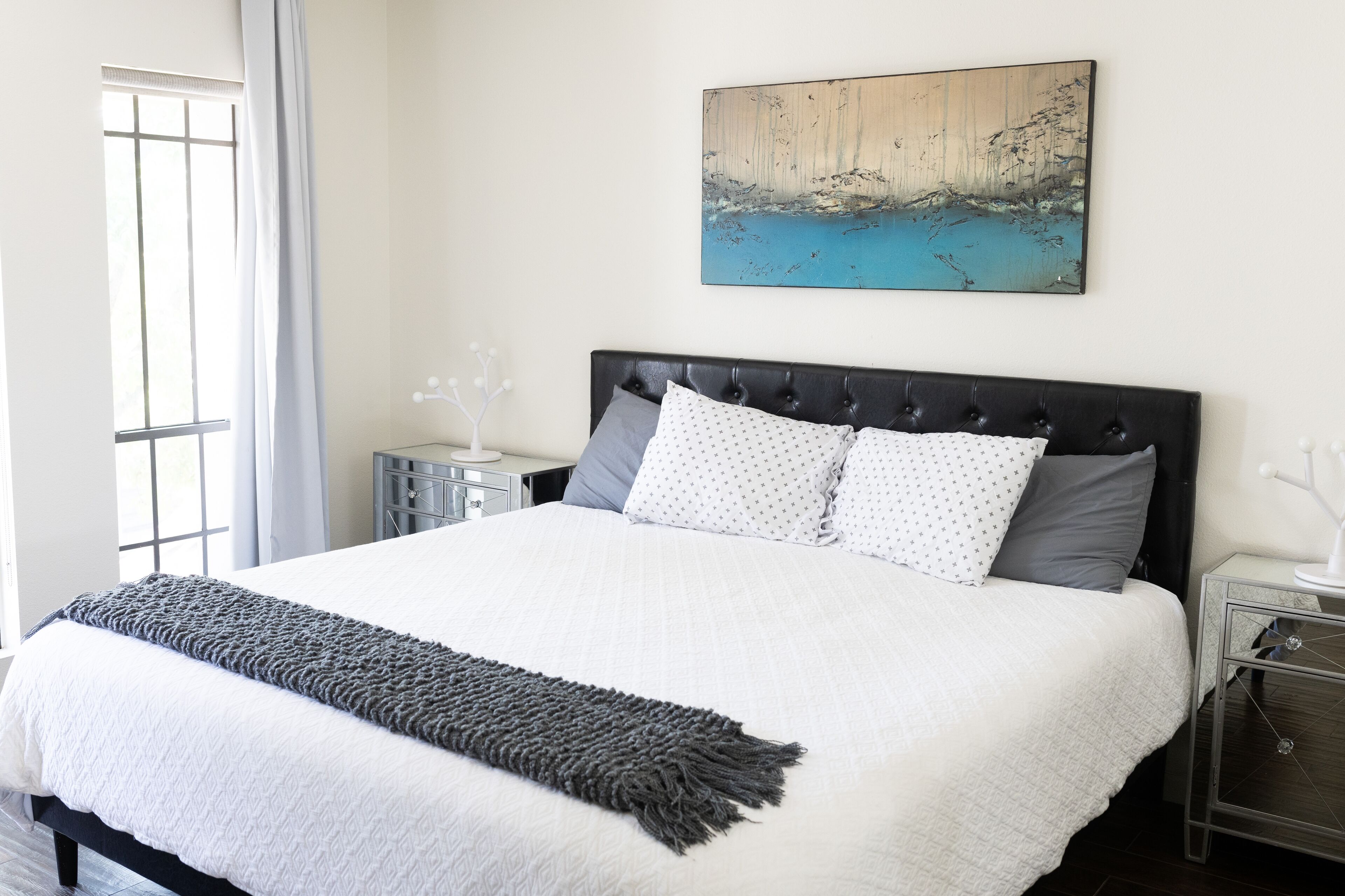 Una cama bien tendida con edredón blanco y manta gris, cabecera tapizada en negro y juego de almohadas gris y blanco.