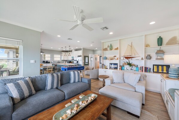 A spacious living area with a gray sofa, a wooden coffee table, and a white armchair. A kitchen with a foosball table is visible in the background.