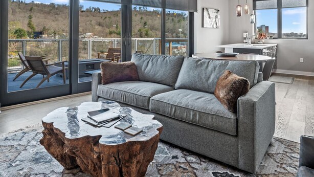 Een moderne woonkamer met een grijze bank, een houten salontafel en grote ramen met uitzicht op een heuvelachtig landschap.