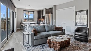 Een moderne woonkamer met een grijze bank, een salontafel en een leren fauteuil. Achtergrond toont een keuken met donkere kastjes en roestvrijstalen apparatuur.