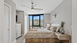 Een slaapkamer met een bed, nachtkastje, kast, plafondventilator en een groot raam met uitzicht op de omgeving.