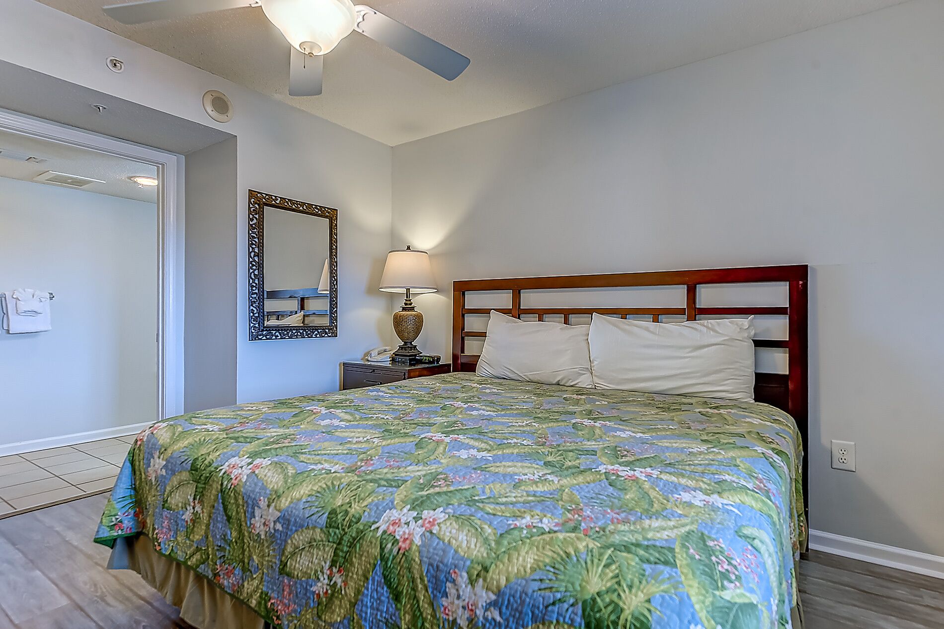 A bedroom with a bed, a nightstand, a lamp, a mirror, and a ceiling fan.