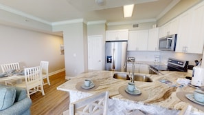 A modern kitchen with white cabinetry, a stainless steel refrigerator, a built-in oven, and a microwave. There is a dining area with a table and chairs.
