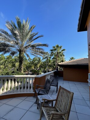A balcony with a tiled floor, two wooden chairs, a white balustrade, and palm trees in the background.