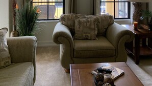 Uma sala de estar aconchegante com sofá, poltrona e mesa de centro. Há duas janelas com cortinas, uma luminária e um vaso com flores.