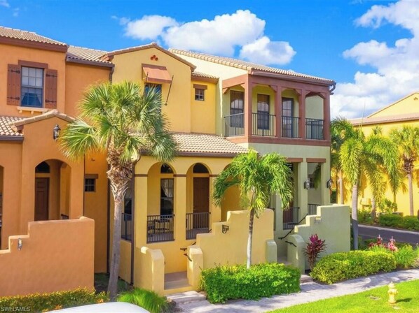 A multi-story residential building with a yellow facade, multiple balconies, and a well-maintained garden with palm trees and shrubs. -  Resort home, walk to it all, swim in two lush resort pools. (Naples)