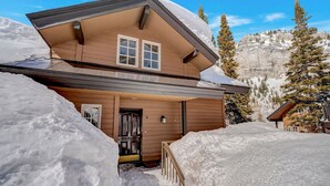A two-story wooden cabin with a sloped roof, surrounded by snow and mountains.