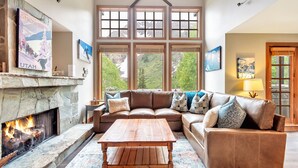 A spacious living room with a stone fireplace, a large window with multiple panes, a leather sectional sofa, and a wooden coffee table.