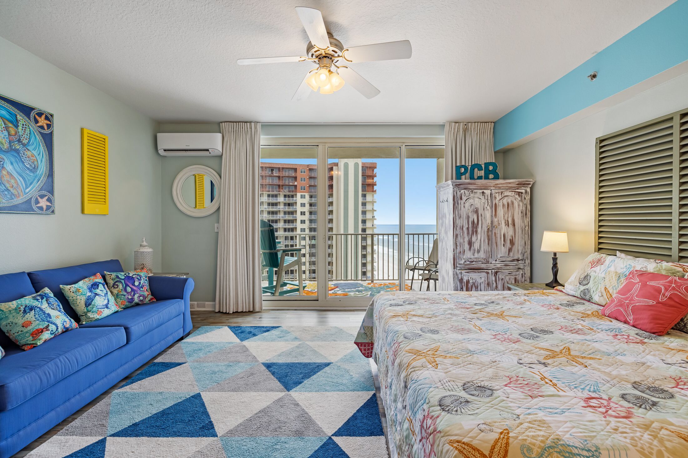 Une chambre d’hôtel avec un canapé bleu, un lit recouvert d’une courtepointe aux couleurs vives, un ventilateur de plafond et une vue sur les immeubles de grande hauteur, visible à travers la porte vitrée.