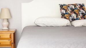 Un lit avec une couverture matelassée grise, un oreiller blanc et un oreiller coloré orné de motifs de chiens. On y trouve une table de chevet en bois avec une lampe et un tiroir.