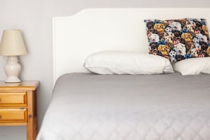 Een bed met een geruite grijze deken, een wit kussen en een kleurrijk kussen met hondjesprintjes. Er staat een houten nachtkastje met een lamp en een lade.