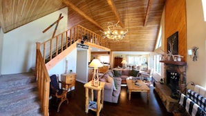 Interior acogedor de una cabaña con una escalera de madera, una sala de estar con sofá y mesa, y una chimenea.