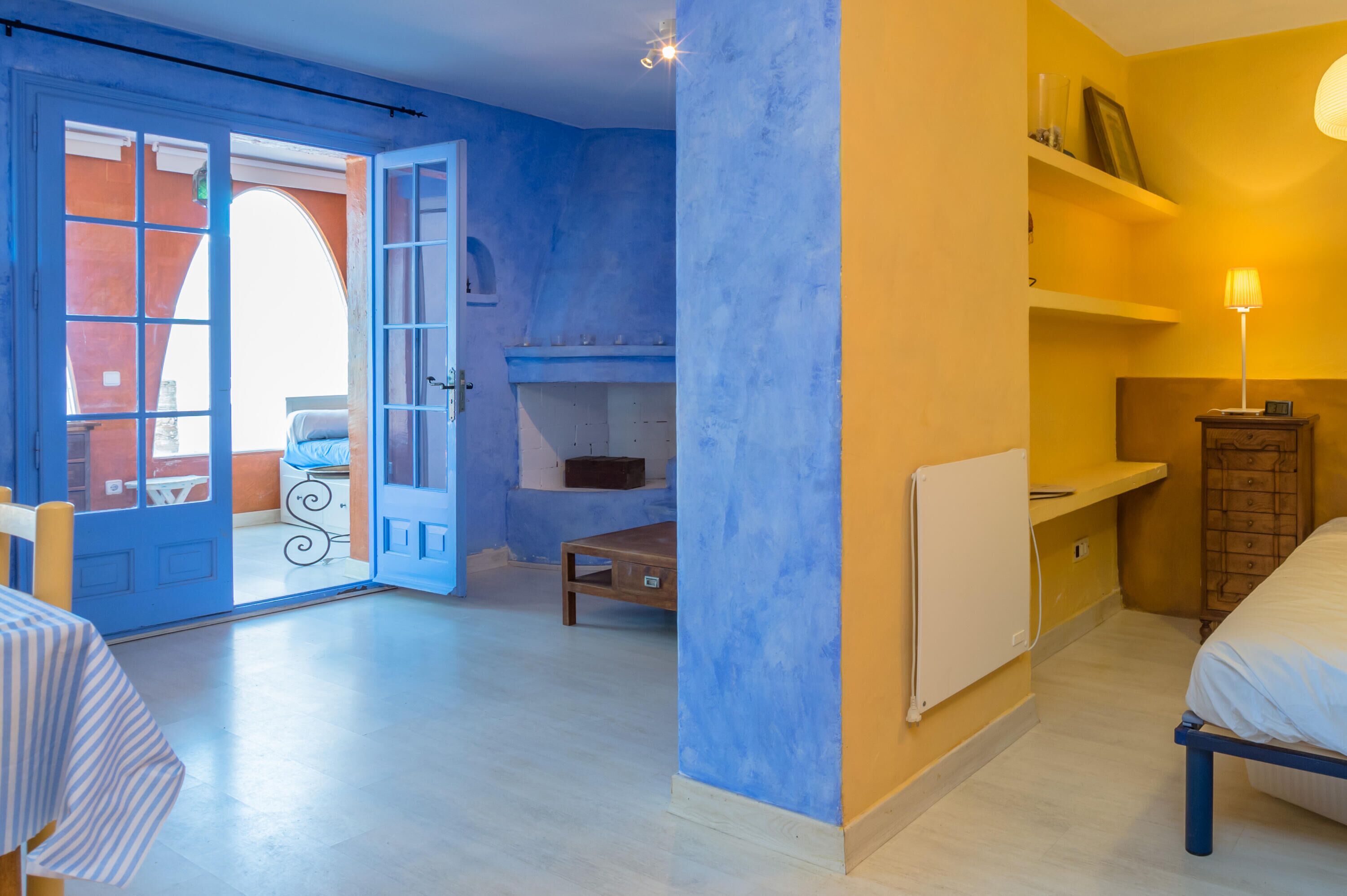 A room with a bed, a table, a chair, a small table, a radiator, and a shelf with items.