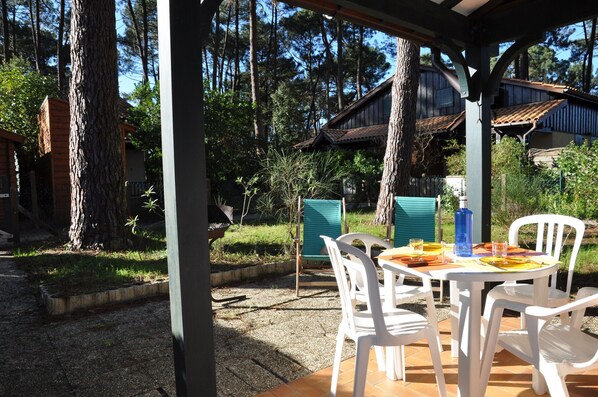 A covered outdoor dining area with white plastic chairs and a table set for two, surrounded by trees and a wooden building in the background.