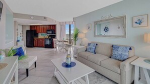 A modern living room with a beige sofa, a coffee table, and a dining area with a table and chairs. A kitchen with dark cabinets and a black refrigerator is visible in the background. - Stunning Oceanview Penthouse Dogs OK (Myrtle Beach)