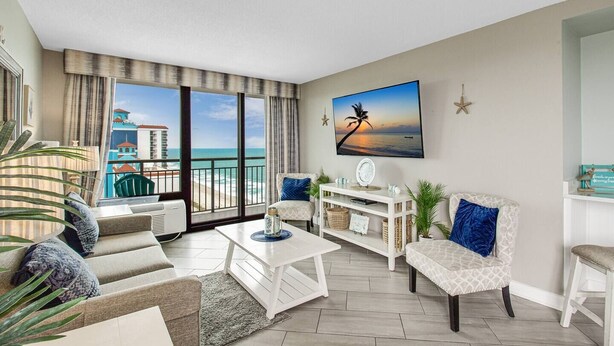 A spacious living room with a large window overlooking a beach, a flat-screen TV mounted on the wall, and a white console table with decorative items. - Stunning Oceanview Penthouse Dogs OK (Myrtle Beach)